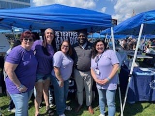 People in purple shirts posing with a man in a suit at an outdoor rally.