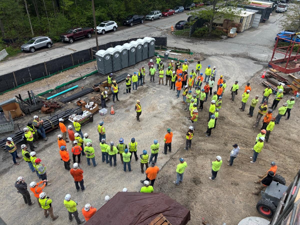 Dozens of construction workers in hard hats and safety vests standing in a large circle at a jobsite, listening to a speaker in the middle