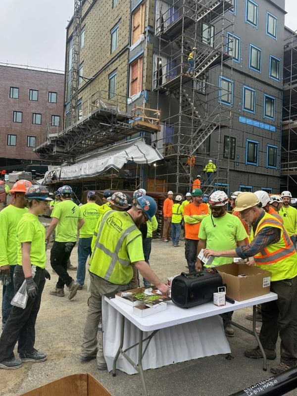Construction workers in safety vests and hard hats at a jobsite table, picking up kits